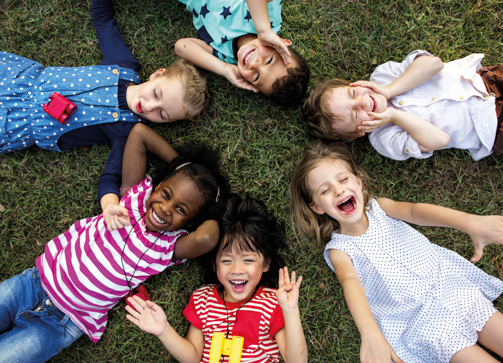 verschiedene Kinder liegen im Kreis auf Rasen, Köpfe in der Mitte, und lachen